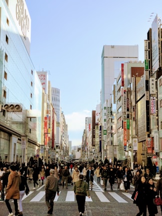 A busy urban street scene with numerous people walking across a wide crosswalk surrounded by tall buildings adorned with advertisements and signs. The clear sky above provides a backdrop that contrasts with the bustling activity below.
