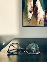A pair of eyeglasses resting on a dark wooden surface in a well-lit room. In the background, a colorful abstract painting with geometric patterns hangs on a white wall. The sunlight casts soft shadows across the table.