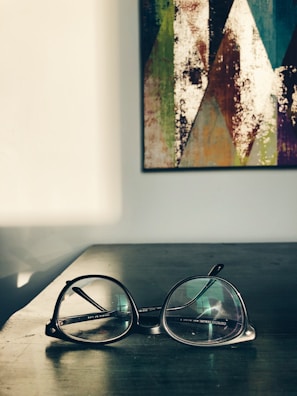A pair of eyeglasses resting on a dark wooden surface in a well-lit room. In the background, a colorful abstract painting with geometric patterns hangs on a white wall. The sunlight casts soft shadows across the table.