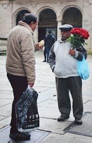 Two men are engaged in conversation on a stone-paved street in front of a building with arches. One man is holding a black plastic bag and what appears to be a sandwich, while the other is holding a bouquet of red roses inside a blue plastic bag. A person in a dark jacket walks in the background.