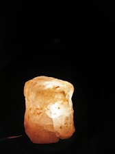 A large, translucent rock salt lamp is illuminated from within, casting a warm and soothing glow. The surface is rough and the color ranges from white to pale orange with scattered darker spots.