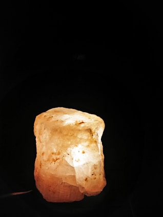 A large, translucent rock salt lamp is illuminated from within, casting a warm and soothing glow. The surface is rough and the color ranges from white to pale orange with scattered darker spots.