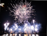An expansive display of fireworks lights up the night sky over a body of water. The fireworks burst in various directions, creating intricate patterns of light and smoke. The reflection of the fireworks can be seen on the water's surface, adding to the visual spectacle.