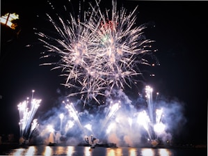An expansive display of fireworks lights up the night sky over a body of water. The fireworks burst in various directions, creating intricate patterns of light and smoke. The reflection of the fireworks can be seen on the water's surface, adding to the visual spectacle.