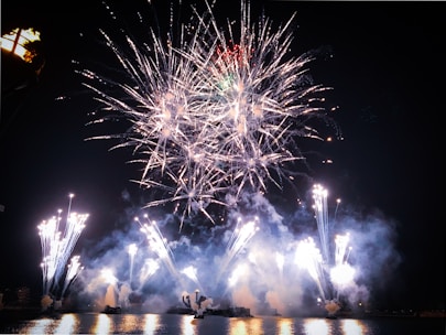 An expansive display of fireworks lights up the night sky over a body of water. The fireworks burst in various directions, creating intricate patterns of light and smoke. The reflection of the fireworks can be seen on the water's surface, adding to the visual spectacle.