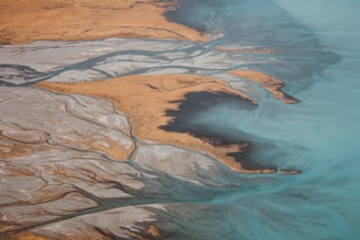 An aerial view of a winding river delta with sandy banks contrasting against expansive turquoise water. The landscape features intricate patterns formed by water channels and land, creating a natural mosaic of earthy tones and light blues.