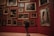 A person stands in an art gallery, facing a wall adorned with numerous framed paintings. The artworks feature various depictions, predominantly of nude figures, and are arranged closely together on a deep red background. The large ornate frames contribute to a classic and refined aesthetic.