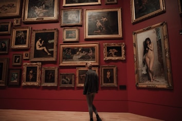 A person stands in an art gallery, facing a wall adorned with numerous framed paintings. The artworks feature various depictions, predominantly of nude figures, and are arranged closely together on a deep red background. The large ornate frames contribute to a classic and refined aesthetic.