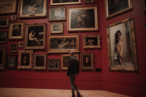 A person stands in an art gallery, facing a wall adorned with numerous framed paintings. The artworks feature various depictions, predominantly of nude figures, and are arranged closely together on a deep red background. The large ornate frames contribute to a classic and refined aesthetic.