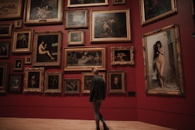 A person stands in an art gallery, facing a wall adorned with numerous framed paintings. The artworks feature various depictions, predominantly of nude figures, and are arranged closely together on a deep red background. The large ornate frames contribute to a classic and refined aesthetic.