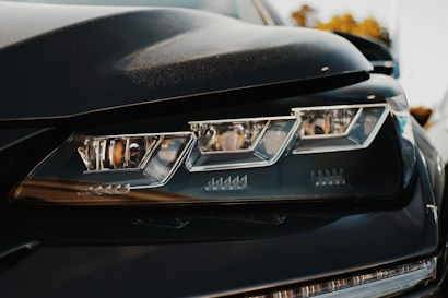 Close-up view of an automobile headlight with a sleek and modern design, showing intricate detailing and sharp angles. The focus is on the reflective surfaces and the geometric shapes of the light elements.