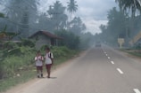 Two children wearing school uniforms walk along a rural road, surrounded by lush vegetation and palm trees. A small house with a rusted roof is visible nearby, and there is a car driving away in the distance under an overcast sky.