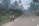 Two children wearing school uniforms walk along a rural road, surrounded by lush vegetation and palm trees. A small house with a rusted roof is visible nearby, and there is a car driving away in the distance under an overcast sky.