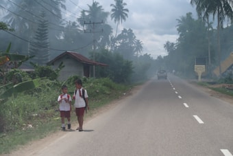 Two children wearing school uniforms walk along a rural road, surrounded by lush vegetation and palm trees. A small house with a rusted roof is visible nearby, and there is a car driving away in the distance under an overcast sky.