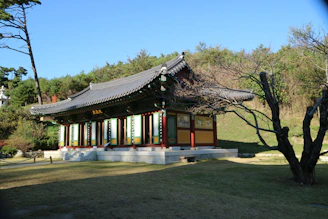 A traditional Korean building with a curved, tiled roof and vibrant painted details sits in a serene garden setting. Surrounding the structure are manicured lawns and neatly trimmed trees, with a backdrop of lush greenery and a clear blue sky.