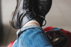 A close-up view of folded blue denim jeans and black leather boots with light stitching details. The jeans have a rolled-up hem, and a white sock peeks out above the boot. The person is resting their leg on a red textured surface with some dark straps or handles visible in the background.