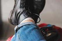 A close-up view of folded blue denim jeans and black leather boots with light stitching details. The jeans have a rolled-up hem, and a white sock peeks out above the boot. The person is resting their leg on a red textured surface with some dark straps or handles visible in the background.