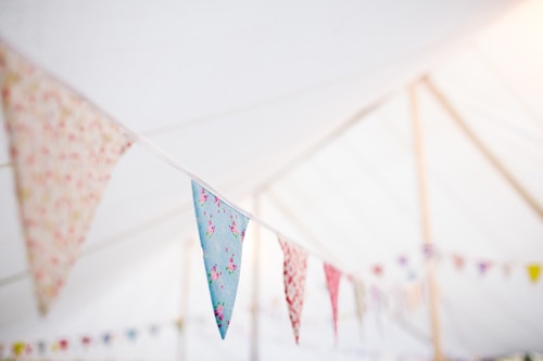 Colorful fabric bunting in a soft-focus setting, strung under a light and airy tent ceiling. The bunting triangles display various patterns, including floral designs.