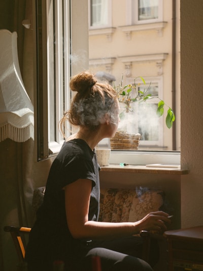 Une femme avec un chignon fume une cigarette assise devant une fenêtre ouverte