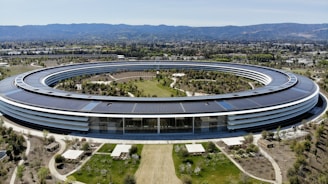 A large, circular building set in a landscaped area with pathways and greenery. The structure features a modern design with extensive use of glass and solar panels on the roof. It is surrounded by well-maintained gardens, trees, and open spaces. In the background, mountains are visible under a clear blue sky.