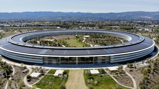 A large, circular building set in a landscaped area with pathways and greenery. The structure features a modern design with extensive use of glass and solar panels on the roof. It is surrounded by well-maintained gardens, trees, and open spaces. In the background, mountains are visible under a clear blue sky.