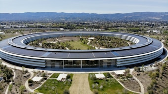 A large, circular building set in a landscaped area with pathways and greenery. The structure features a modern design with extensive use of glass and solar panels on the roof. It is surrounded by well-maintained gardens, trees, and open spaces. In the background, mountains are visible under a clear blue sky.