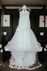 A white wedding dress with lace details is hanging on a wooden frame in a room. The dress has a full tulle skirt and intricate lace bodice, creating an elegant and romantic appearance.