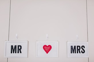 Three framed signs are hanging on a wall. The left sign says 'MR,' the middle sign has a red heart, and the right sign says 'MRS.' Each sign is in a white frame with black text, except for the red heart.