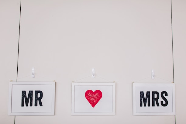 Three framed signs are hanging on a wall. The left sign says 'MR,' the middle sign has a red heart, and the right sign says 'MRS.' Each sign is in a white frame with black text, except for the red heart.