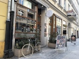 A charming urban street features a café with large glass windows adorned with handwritten menu items. Potted plants and a bike are placed in front of the café, adding a cozy feel. Two sandwich boards display colorful food images and specials. The street is lined with historic European buildings, and a few pigeons are visible on the cobblestone pavement.