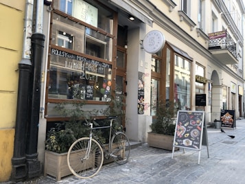 A charming urban street features a caf&eacute; with large glass windows adorned with handwritten menu items. Potted plants and a bike are placed in front of the caf&eacute;, adding a cozy feel. Two sandwich boards display colorful food images and specials. The street is lined with historic European buildings, and a few pigeons are visible on the cobblestone pavement.