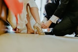 A person is kneeling on the ground, helping another person adjust a pair of gold high-heeled shoes. The scene suggests an intimate or special occasion, as the person receiving assistance is dressed in a white gown, partially visible in the image.