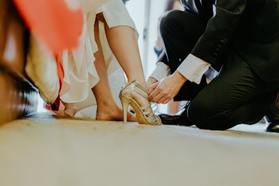 A person is kneeling on the ground, helping another person adjust a pair of gold high-heeled shoes. The scene suggests an intimate or special occasion, as the person receiving assistance is dressed in a white gown, partially visible in the image.