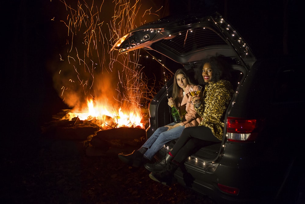 two women sitting on back of SUV near campfire