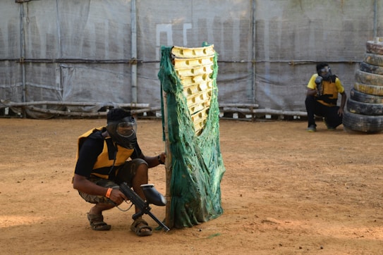 Two individuals are engaged in a paintball game, wearing protective vests and masks. One person is kneeling behind a green and yellow barricade, holding a paintball marker. The second person is crouched behind a stack of tires in the background. The ground is sandy, and there is a large canvas wall enclosing the area.