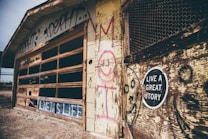 An old, weathered garage with broken windows, covered in graffiti and faded paint. The wooden exterior is deteriorating and features a sign with the words 'Live a Great Story'. The ground is barren with scattered debris.