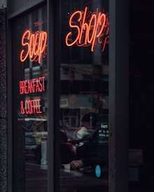 A dimly lit storefront window features neon signs displaying the words 'Shop' and 'Breakfast & Coffee.' Inside, a person can be seen holding a white cup amidst a cozy atmosphere filled with various items on display.