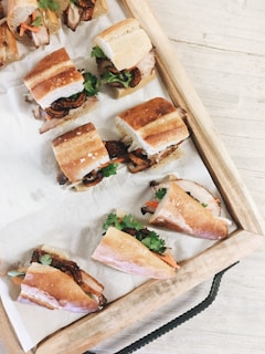 A wooden tray holds several pieces of sandwiches filled with ingredients like cucumber slices, cilantro, meat, and carrots. The sandwiches are arranged neatly on a piece of parchment paper, showcasing various colors and textures of the fillings.