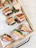 A wooden tray holds several pieces of sandwiches filled with ingredients like cucumber slices, cilantro, meat, and carrots. The sandwiches are arranged neatly on a piece of parchment paper, showcasing various colors and textures of the fillings.