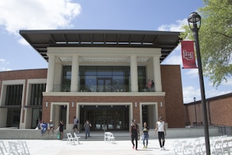 A modern building with large pillars and glass windows, surrounded by a brick exterior. Several people are walking and sitting outside, enjoying a sunny day. A red flag with an insignia is attached to a nearby lamppost. The sky is clear with a few white clouds, and there are green trees providing some shade.