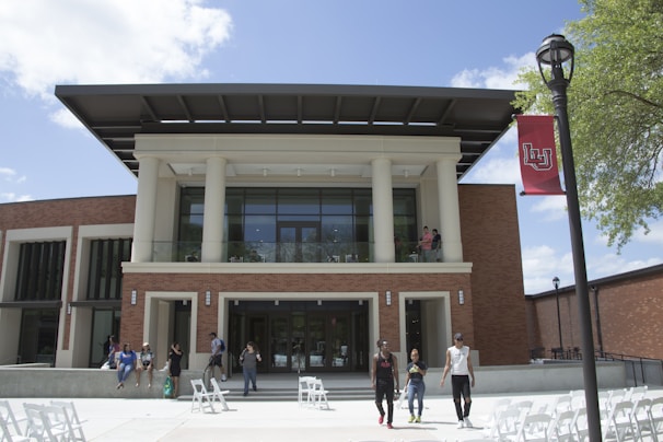 A modern building with large pillars and glass windows, surrounded by a brick exterior. Several people are walking and sitting outside, enjoying a sunny day. A red flag with an insignia is attached to a nearby lamppost. The sky is clear with a few white clouds, and there are green trees providing some shade.