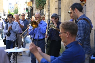 A group of musicians playing instruments on a city street, including a trombone and a clarinet. Several people are gathered around, some clapping along and others watching the performance. The scene appears to be in an urban area with buildings and a pedestrian walkway visible in the background.