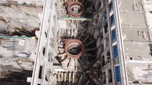 An aerial view of a large industrial or construction site with prominent circular structures and various building materials scattered around. The site is bordered by walls and encompasses machinery designed for heavy-duty work. Details include metallic and concrete textures, with a network of platforms and scaffolding indicating ongoing construction or maintenance activity.