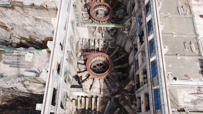 An aerial view of a large industrial or construction site with prominent circular structures and various building materials scattered around. The site is bordered by walls and encompasses machinery designed for heavy-duty work. Details include metallic and concrete textures, with a network of platforms and scaffolding indicating ongoing construction or maintenance activity.