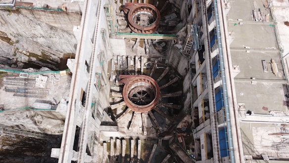 An aerial view of a large industrial or construction site with prominent circular structures and various building materials scattered around. The site is bordered by walls and encompasses machinery designed for heavy-duty work. Details include metallic and concrete textures, with a network of platforms and scaffolding indicating ongoing construction or maintenance activity.