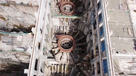 An aerial view of a large industrial or construction site with prominent circular structures and various building materials scattered around. The site is bordered by walls and encompasses machinery designed for heavy-duty work. Details include metallic and concrete textures, with a network of platforms and scaffolding indicating ongoing construction or maintenance activity.
