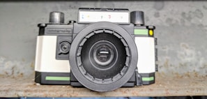 A vintage-style camera with a prominent lens, featuring black, white, and green colors. The camera is placed on a rustic, textured surface with a blurred background.
