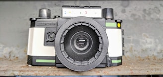 A vintage-style camera with a prominent lens, featuring black, white, and green colors. The camera is placed on a rustic, textured surface with a blurred background.