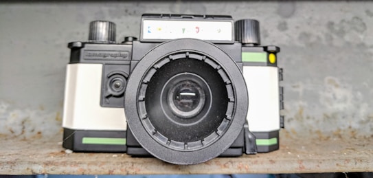 A vintage-style camera with a prominent lens, featuring black, white, and green colors. The camera is placed on a rustic, textured surface with a blurred background.