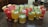 An assortment of colorful beverages is arranged on a granite countertop. The drinks are presented in various types of glassware and garnished with fresh fruits such as watermelon slices, lime wedges, and mint leaves. Ice cubes are visible in some of the glasses, suggesting they are refreshing and cold.
